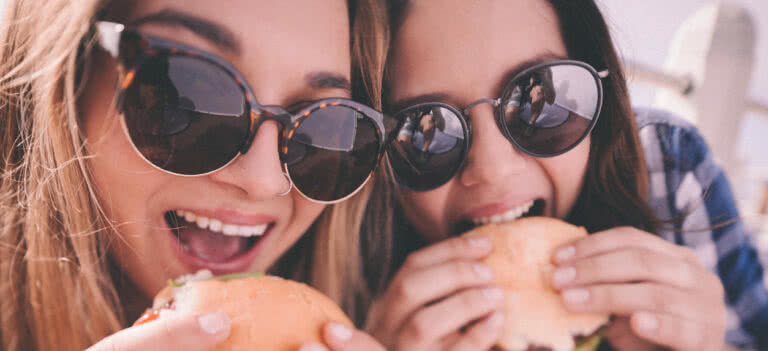 Two women wearing sunglasses posing while each's eating a burger.